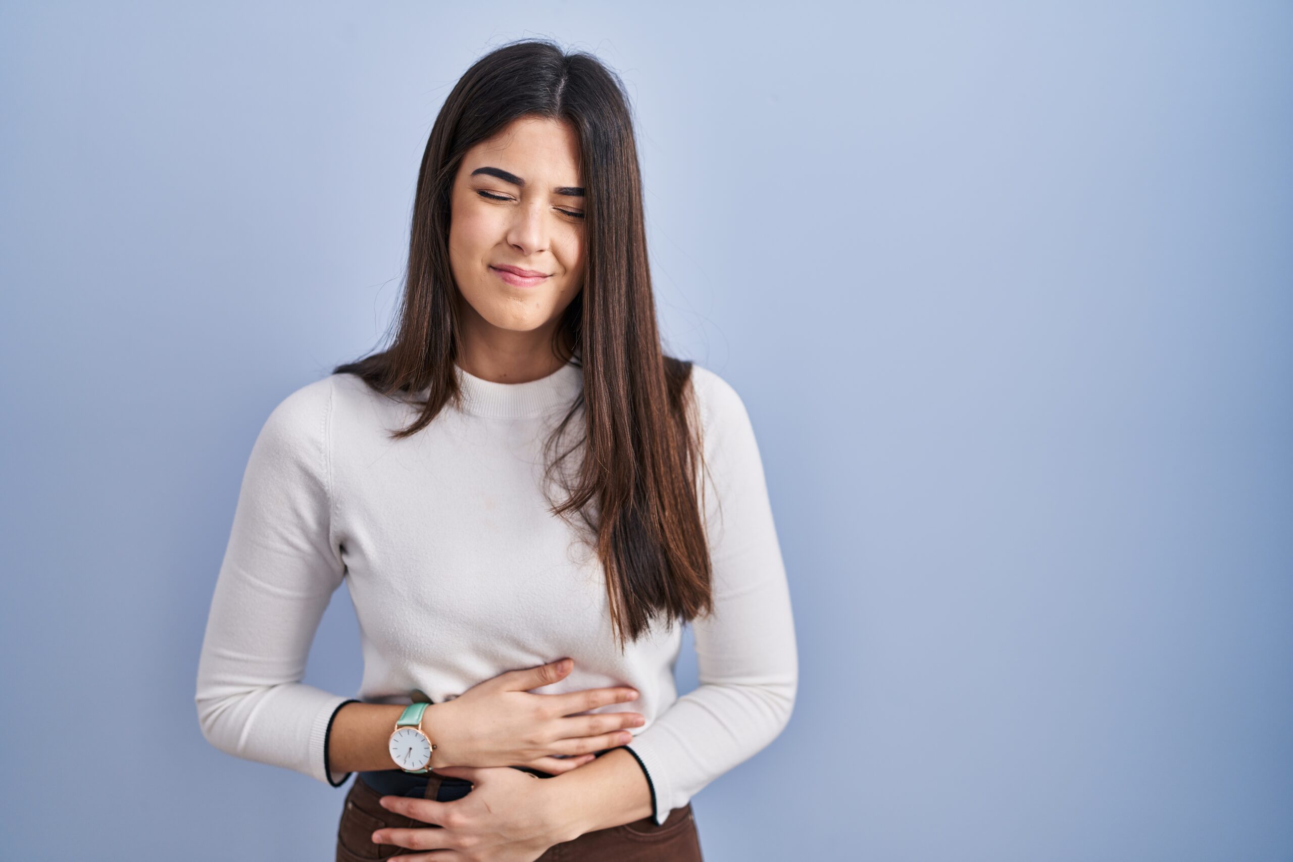 young brunette woman standing blue background with hand on stomach who needs natural wellness